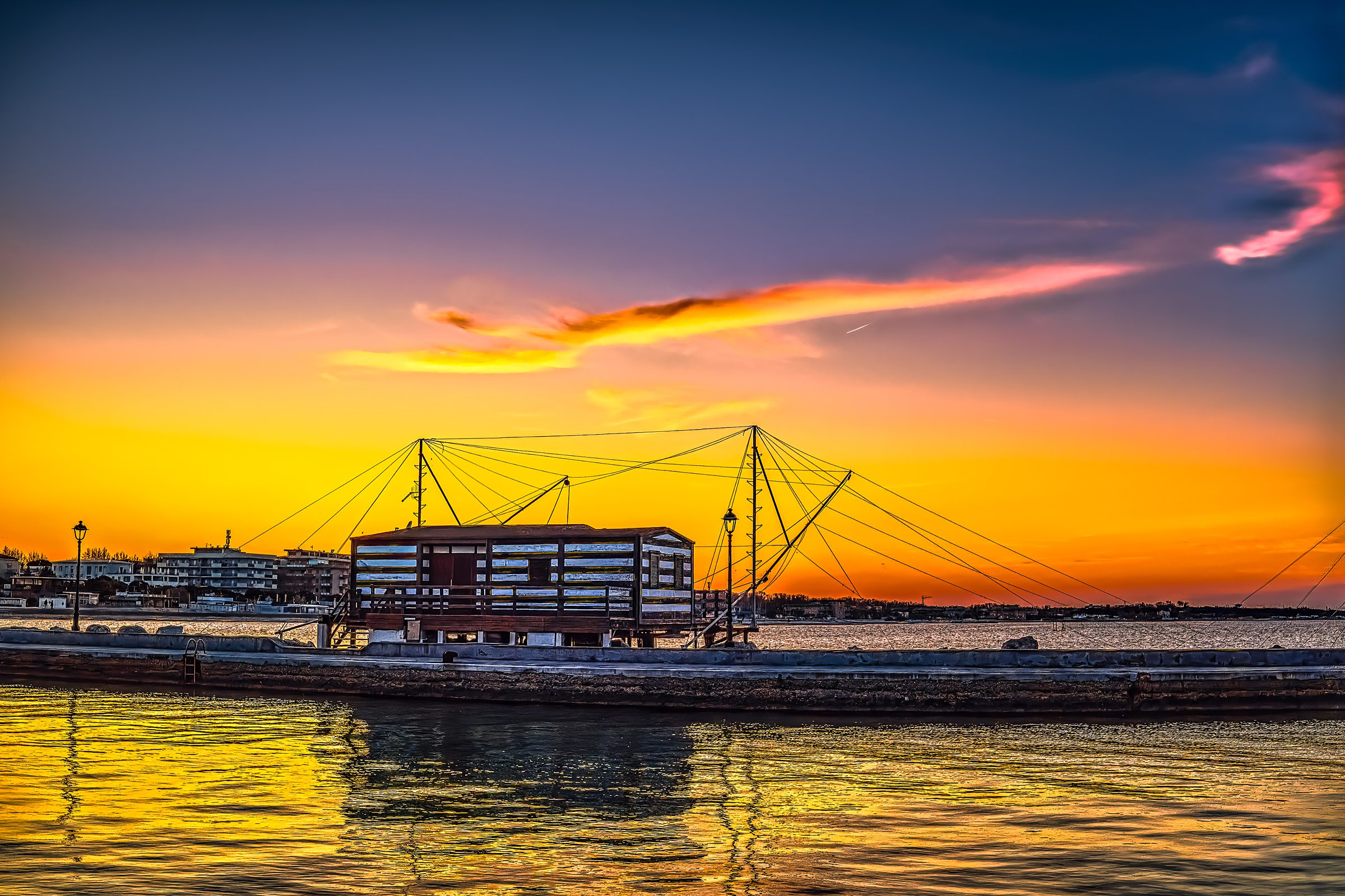 Silhouetted fishing hut in Cesenatico against radiant sunset with golden reflections on still waters