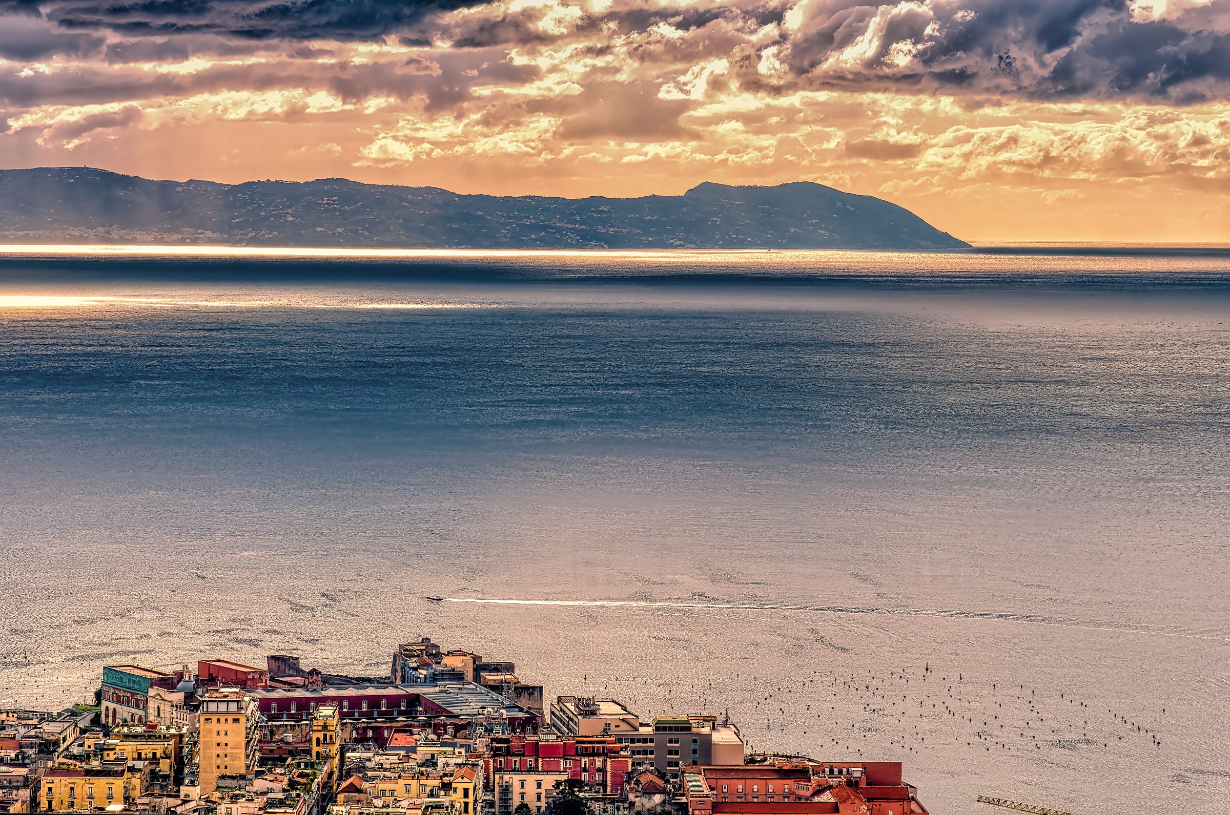 Golden hour sunset over Mediterranean coastal town with colorful buildings cascading to crystal-clear waters, dramatic clouds creating warm reflections